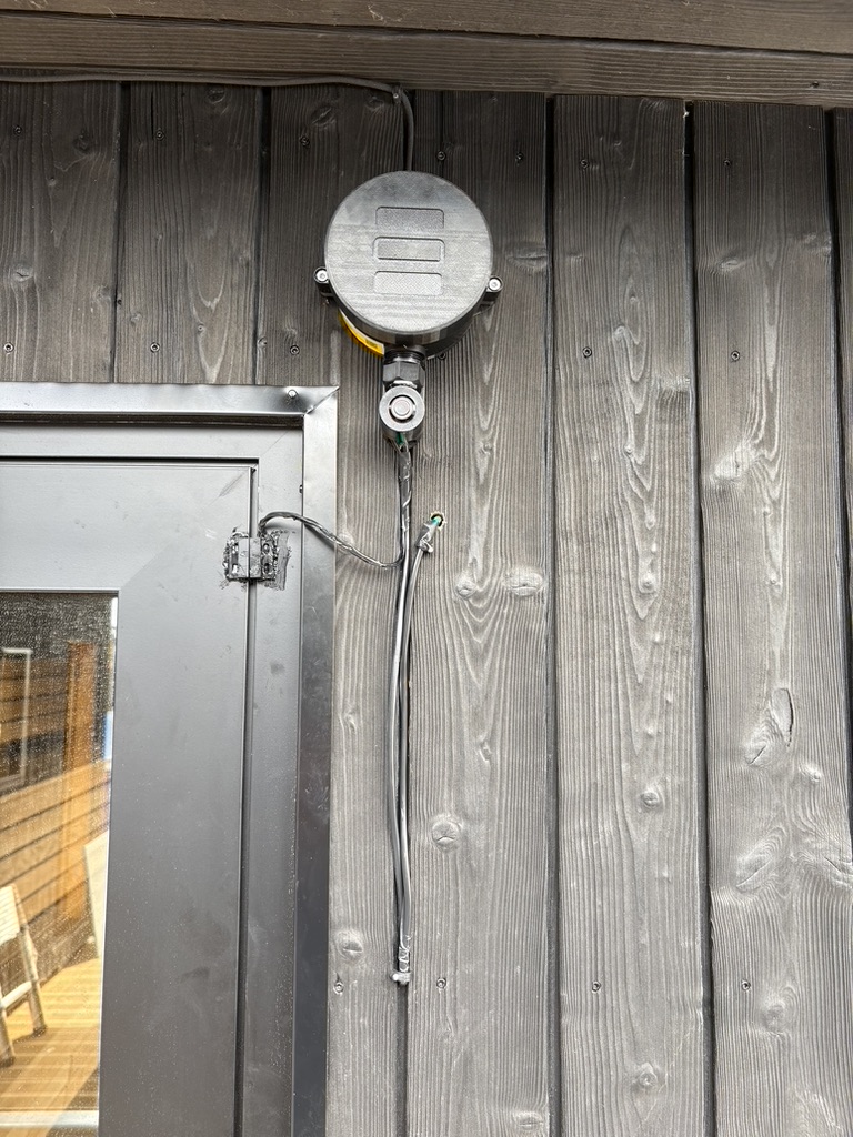 SenseStack Base Device surface-mounted on a timber sauna wall beside the door, with probe and door sensor cabling routed down the frame